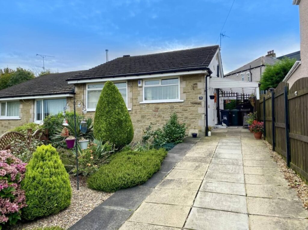A small stone bungalow with a driveway, garden featuring trimmed shrubs and flowers, and a side gate.
